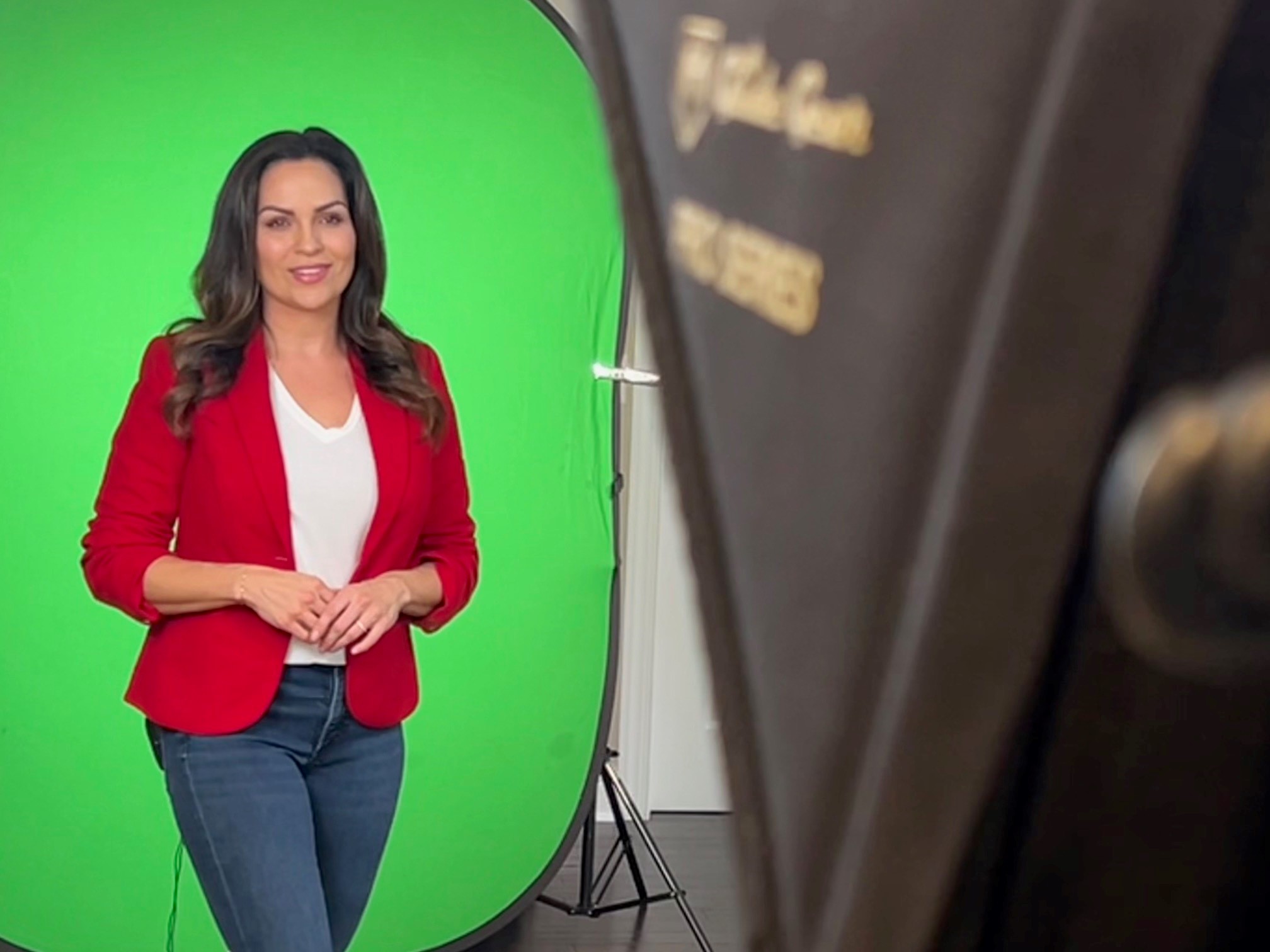 A lady with brunette hair smiling in front of a camera with a green screen behind her. She is wearing a red jacket